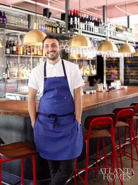 Man in blue apron standing behind a bar with shelves of liquor bottles and brass pendant lights. Red bar stools are visible in the foreground.