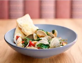 Bowl of clams with spinach, tomatoes, and crusty bread on a wooden table with a blurred background of pink and brown striped fabric.