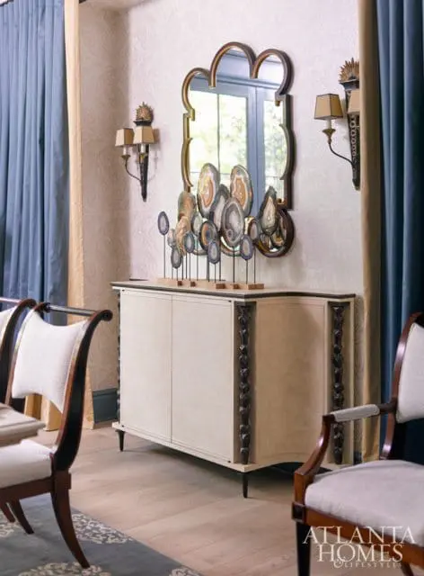 Dining room with a cream-colored cabinet topped with agate slices, a decorative mirror, and sconces on a textured wallpapered wall.