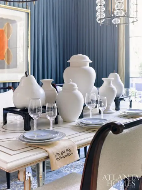 Dining room table set with white decorative vases, a pear sculpture, and place settings. Blue curtains hang in the background.