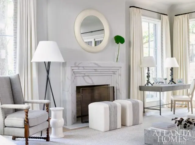 Living room with a gray upholstered armchair, marble fireplace, two white ottomans with gray stripes, and a desk with lamps.