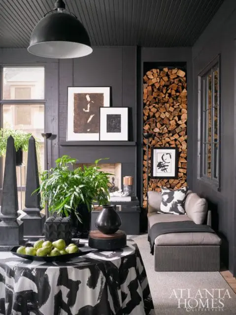 Dark paneled room with a fireplace stacked with wood, a chaise lounge with patterned pillows, and a table covered in a black and white abstract print tablecloth with a bowl of pears.