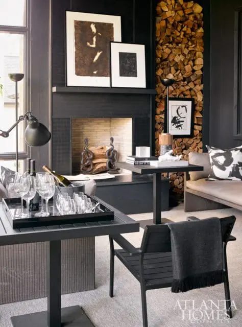 Living room with black fireplace, stacked wood wall, framed art, and a tray with wine glasses and bottles on a dark table.