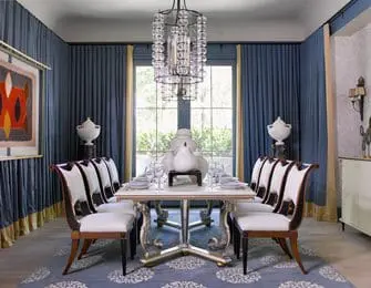 Formal dining room with blue walls, gold-trimmed blue curtains, a rectangular dining table with ornate silver base, white upholstered chairs with dark wood frames, and a crystal chandelier.