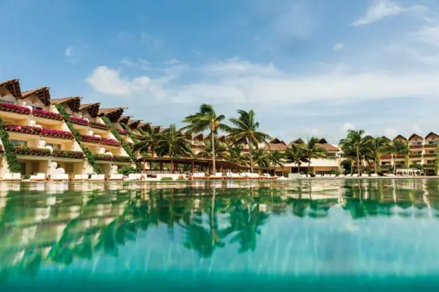 Resort with tiered balconies adorned with flowers and thatched roofs, overlooking a large pool reflecting palm trees and buildings under a blue sky.