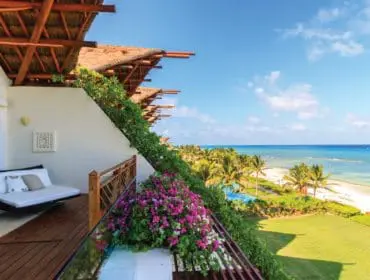 Balcony with thatched roof overhang, white daybed, and lush greenery overlooking a tropical beach with palm trees and turquoise ocean.