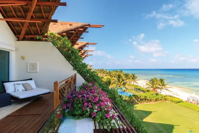 Balcony with thatched roof overhang, white daybed, and lush greenery overlooking a tropical beach with palm trees and turquoise ocean.