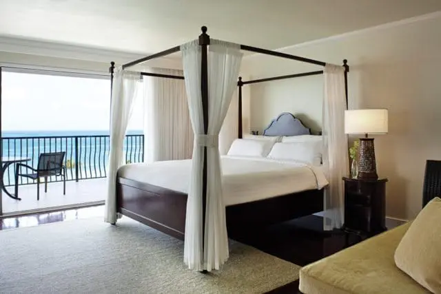 Bedroom with a dark wood four-poster bed draped in white sheer curtains, white bedding, and a blue arched headboard. A balcony with ocean view is visible through sliding glass doors.