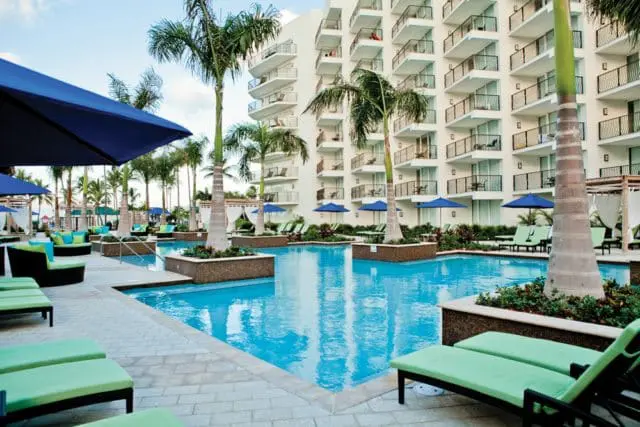 Resort pool area with bright blue water, surrounded by palm trees, lounge chairs, and a multi-story white building with balconies.