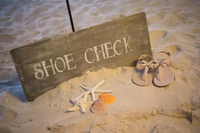 Wooden sign reading 'SHOE CHECK' on a sandy beach with starfish, shells, and a pair of light pink sandals with bows and rhinestones.