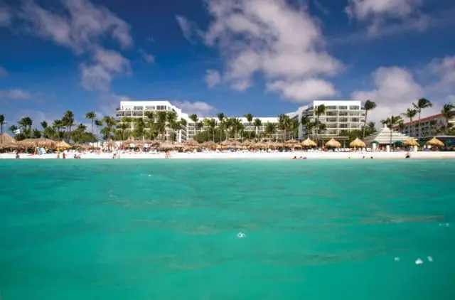 White resort buildings with balconies line a sandy beach with palm trees and thatched umbrellas, viewed from turquoise ocean water.