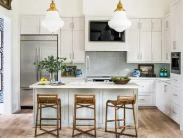 White kitchen with light wood floors, stainless steel refrigerator, island with three bamboo stools, and white cabinetry.