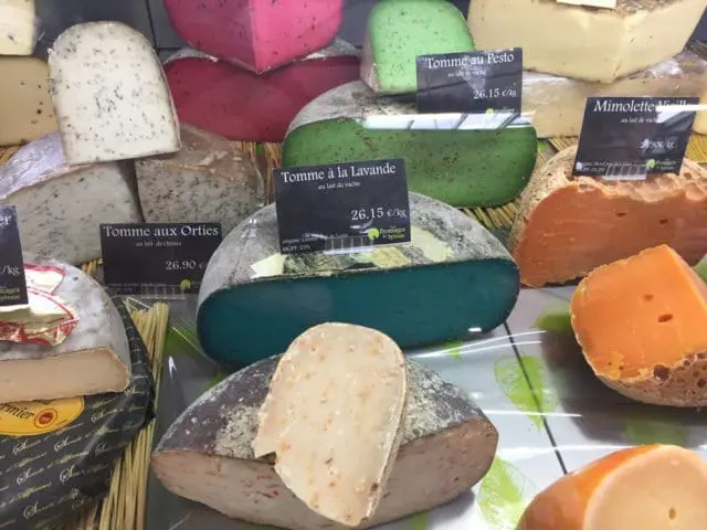 Assortment of artisanal cheeses including Tomme aux Orties, Tomme à la Lavande, Tomme au Pesto, and Mimolette Vieille, with visible price tags.