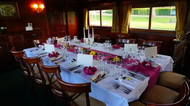 Formal dining room with a long table set for dinner, featuring a white tablecloth, magenta runner, floral centerpieces, and wooden chairs.