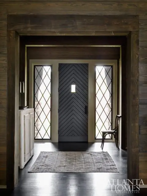 Dark wood paneled entryway with a dark gray chevron door, diamond-paned windows, a distressed white cabinet, and a patterned rug on dark wood floors.
