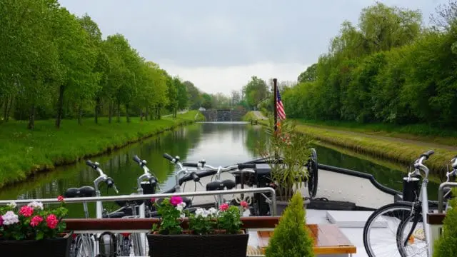 Bicycles parked on the deck of a boat on a canal lined with green trees and grass, with a lock and bridge in the distance.