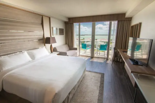 Guest room with king-size bed, upholstered headboard, sofa, desk with television, and balcony overlooking the ocean.