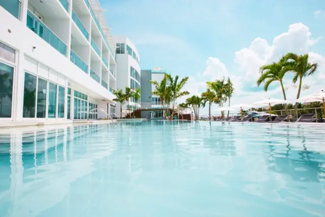 Modern white hotel building with balconies overlooking a bright blue swimming pool and palm trees under a cloudy sky.