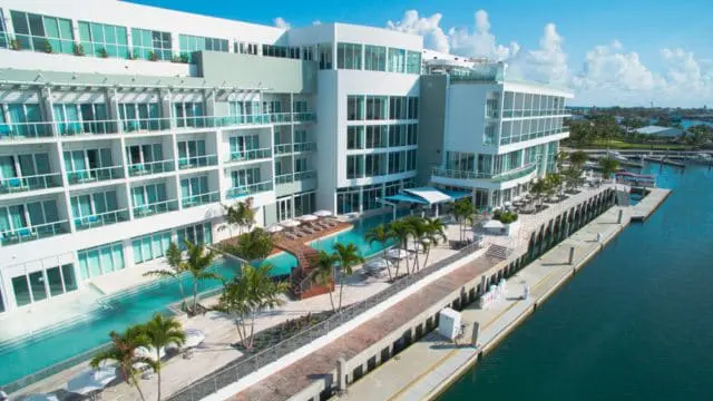 Modern white hotel building with balconies overlooking a lagoon-style pool and marina on a sunny day.