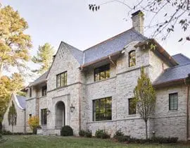 Stone Tudor-style home with dark-framed windows, copper gutters, and a slate roof, surrounded by mature trees and a manicured lawn.