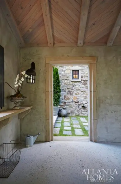 Interior view of a room with a rough plaster wall, wooden beamed ceiling, and a stone doorway leading to a courtyard with a stone wall and stepping stones.