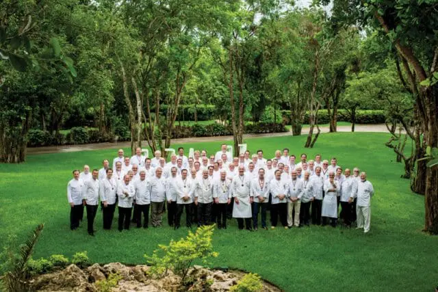A large group of chefs in white uniforms stand together on a lush green lawn in front of a backdrop of trees and foliage.
