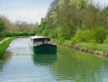 Green barge moving down a canal lined with trees and a walking path on the left. Lush green grass and foliage on both banks.
