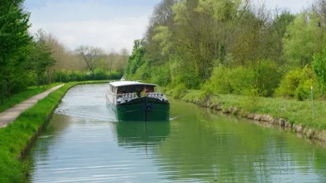 Green barge moving down a canal lined with trees and a walking path on the left. Lush green grass and foliage on both banks.