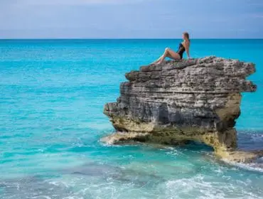 Woman in black swimsuit sitting on a rocky outcrop in turquoise ocean water.