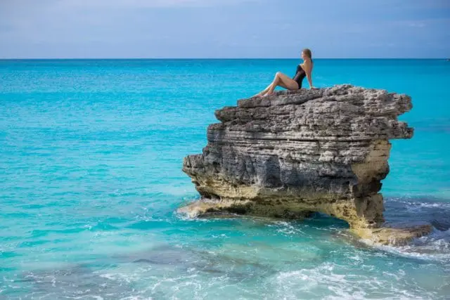Woman in black swimsuit sitting on a rocky outcrop in turquoise ocean water.