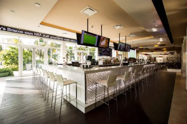 Modern bar with a white marble countertop, white bar stools, and a decorative geometric pattern on the front. Multiple flat-screen TVs are mounted above the bar.