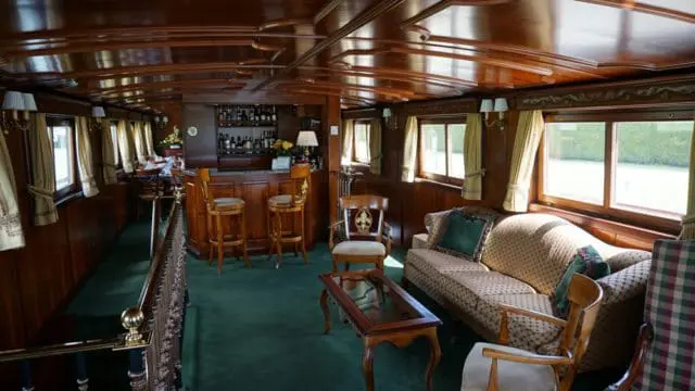 Salon with dark wood paneling, green carpet, a bar with stools, a sofa, armchairs, and a coffee table.
