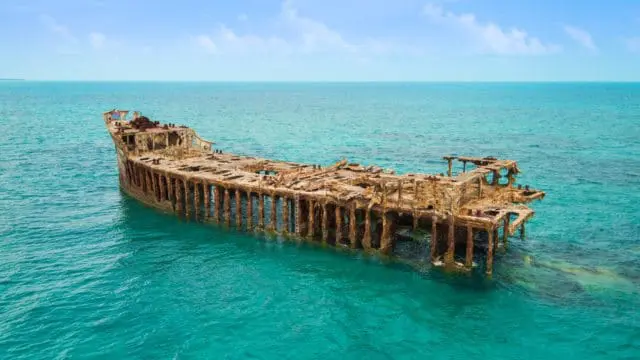 Rusted shipwreck partially submerged in clear turquoise water under a bright blue sky with scattered clouds.