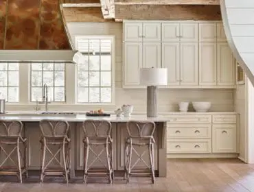 Cream kitchen with a copper range hood, white marble island, and woven bar stools. White cabinetry and wood beam ceiling complete the rustic design.