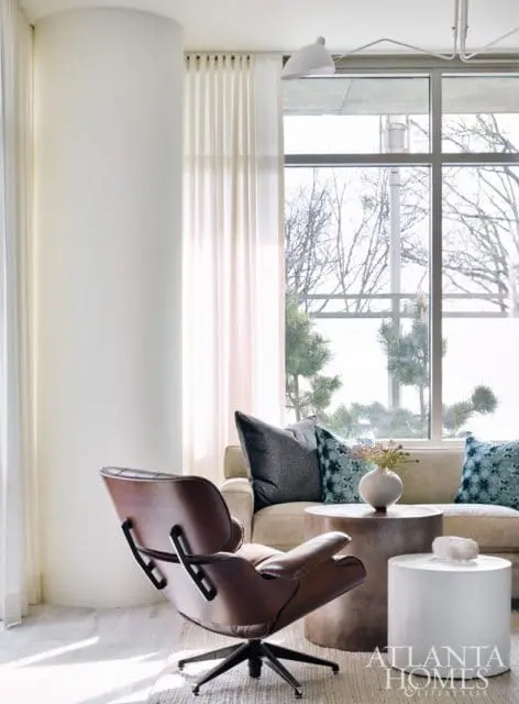 Living room with a brown leather swivel chair, a cream sofa with patterned pillows, and a metal side table holding a vase. A large window overlooks bare trees.
