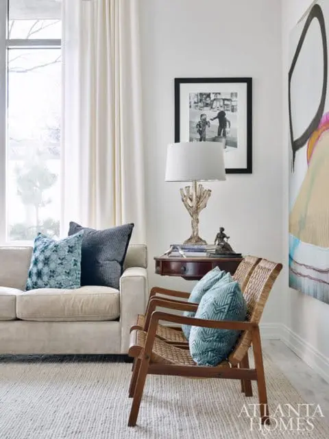 Living room with a cream sofa, two woven armchairs, and a small wooden side table with a lamp and artwork on the wall.