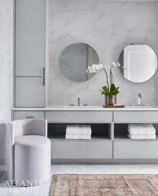 Gray bathroom with marble tile walls, double vanity with white countertops, two round backlit mirrors, and a textured gray swivel stool.
