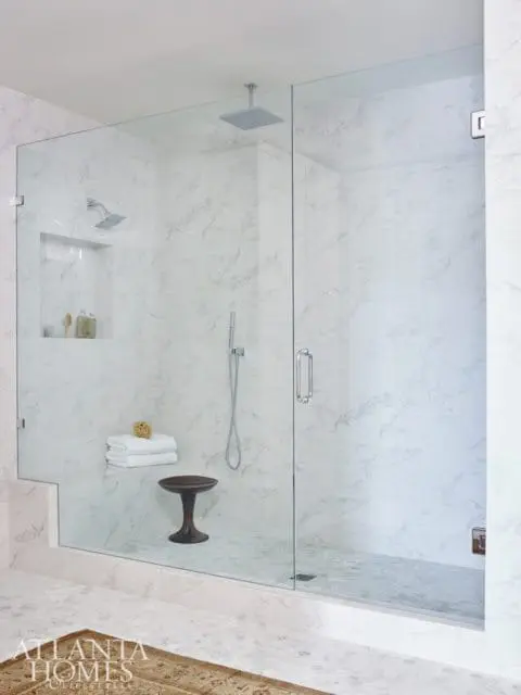 Modern bathroom with a large glass-enclosed shower featuring marble tile walls, a rain shower head, and a built-in bench. A small wooden stool sits inside the shower.