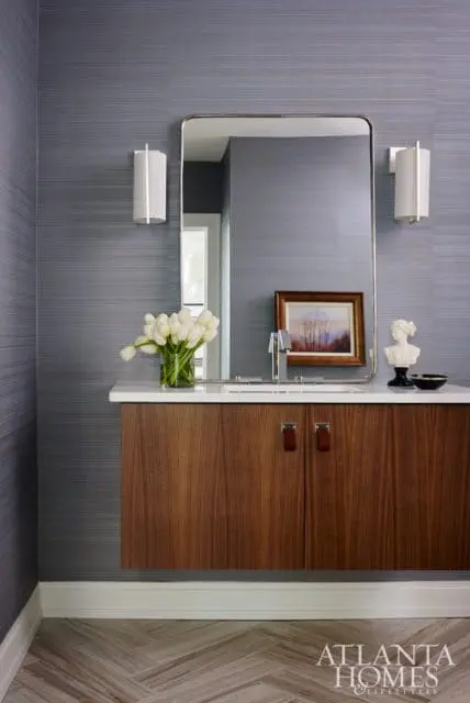 Modern bathroom vanity with dark wood cabinets, white countertop, and a large rectangular mirror. Blue textured wallpaper and sconces adorn the walls.