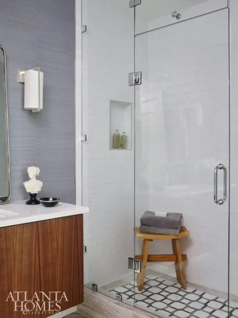 Modern bathroom with a glass-enclosed shower featuring white subway tile walls and a geometric patterned floor. A wooden stool holds folded gray towels. A vanity with a wood grain cabinet and white countertop is visible.