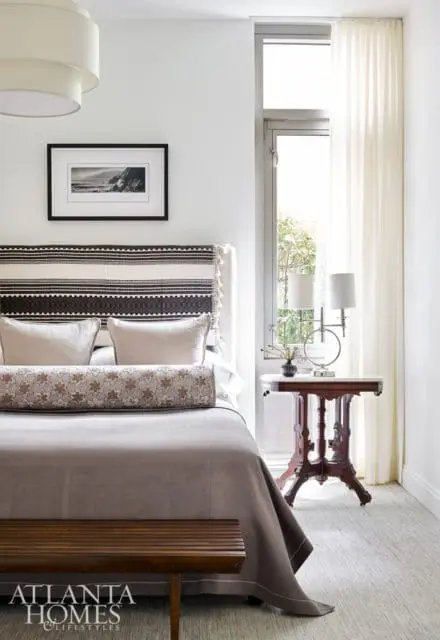 Bedroom with a king-size bed featuring a patterned headboard, neutral linens, and a bolster pillow. A framed black and white photograph hangs above the bed. A small wooden bench sits at the foot of the bed.