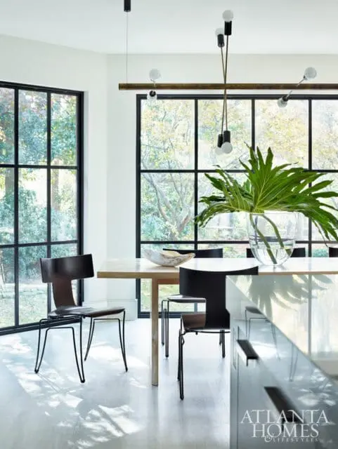 Modern dining room with light wood table, black chairs, large windows overlooking trees, and a contemporary chandelier.