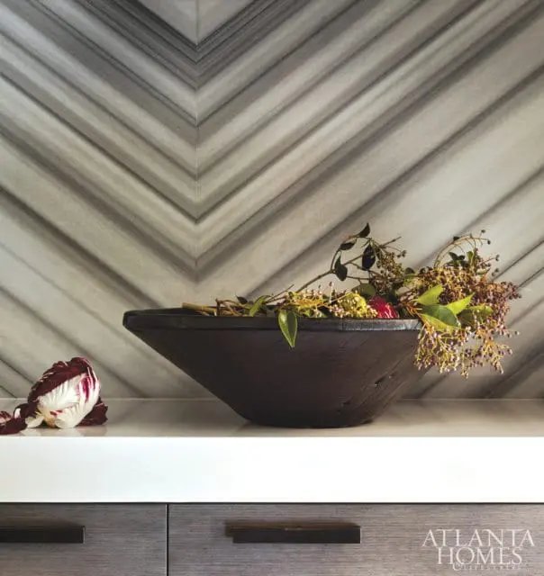 Close-up of a dark wooden bowl filled with branches and berries, placed on a white countertop in front of a chevron-patterned marble backsplash. A radicchio is on the left.