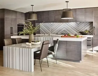 Modern kitchen with gray cabinetry, a marble waterfall island, and a geometric marble backsplash. A dining table with chairs sits in the foreground.