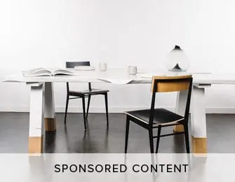 Dining table with white top and white legs accented with gold at the base, set with chairs, books, and vases against a white wall.