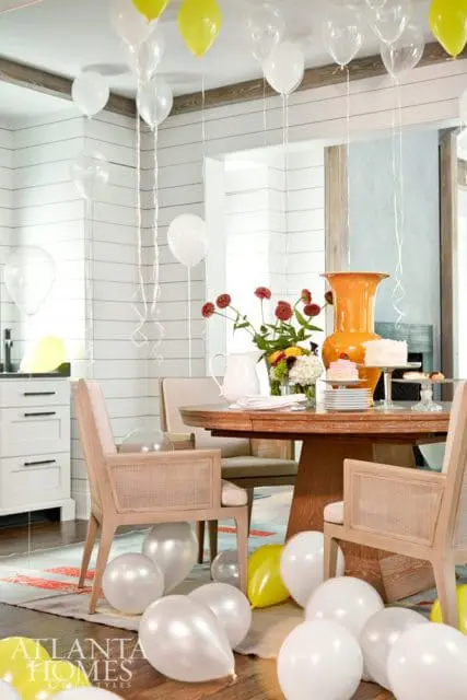 Dining room with a round wooden table set for a celebration, adorned with cakes, flowers, and white and yellow balloons.