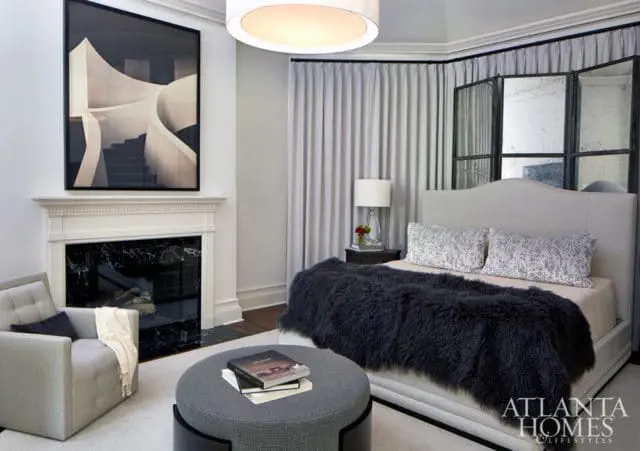 Bedroom with a light gray upholstered bed, dark gray faux fur throw, patterned pillows, a white fireplace with black marble surround, and a large abstract artwork above the fireplace.