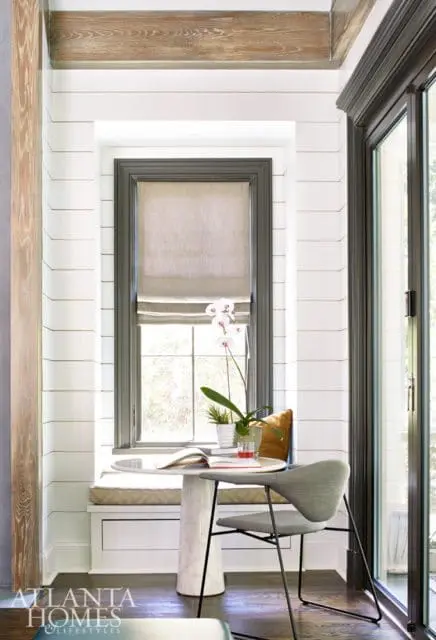 Cozy reading nook with a marble pedestal table, gray upholstered chair, and a window seat with shiplap walls and a linen Roman shade.