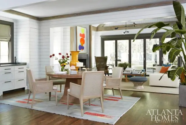 Dining area with a round wooden table and four upholstered chairs, set on a light blue rug with red and yellow accents. White shiplap walls and exposed wood beams accent the space.
