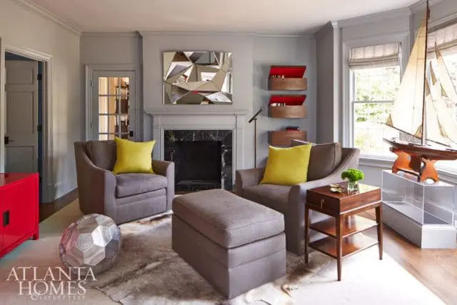 Gray living room with two gray armchairs, a gray ottoman, a fireplace with a marble surround, a geometric mirror, and a model sailboat on a stand.